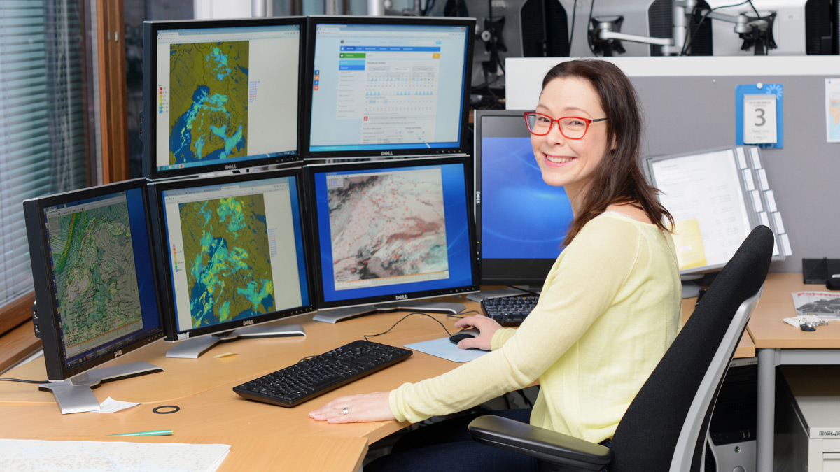 Hymyilevä meteorologi työpisteellään useamman tietokoneen ruudun ääressä.