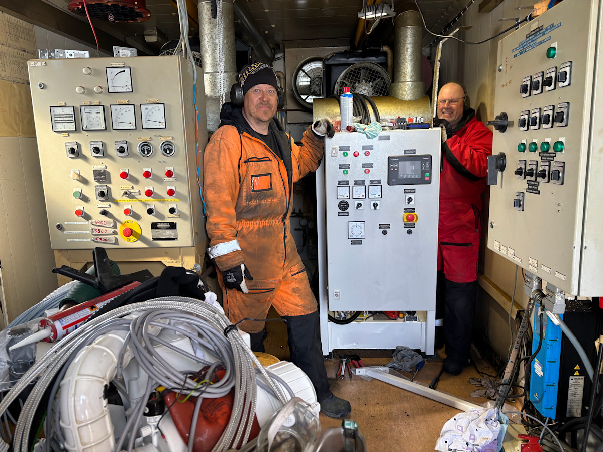 Kaksi miestä työhaalareissa pienessä kontissa laitteiden ja johtojen keskellä.