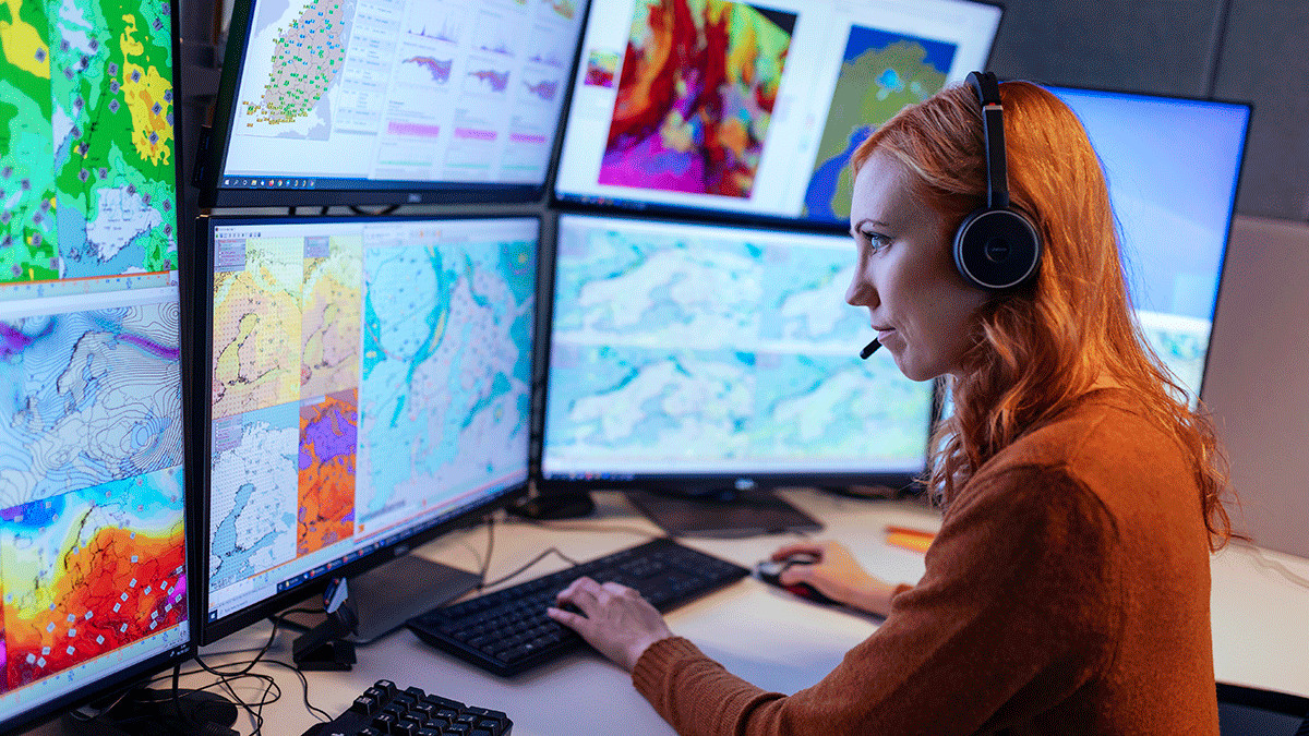 Punahiuksinen naismeteorologi kuulokkeet korvilla usean ison tietokoneruudun ääressä kädet näppäimistöllä. Ruuduilla värikkäitä karttakuvia.