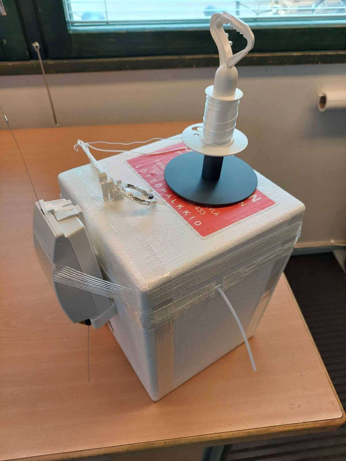 Valkoinen neliön muotoinen laatikko, jossa on kiinni mittalaite sekä lankarulla.