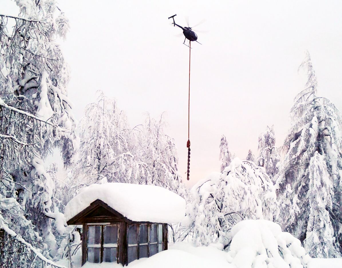 Helikopteri lentää lumisessä maisemassa roikottaen työkalua, joka pudottaa tykkylunta puista.