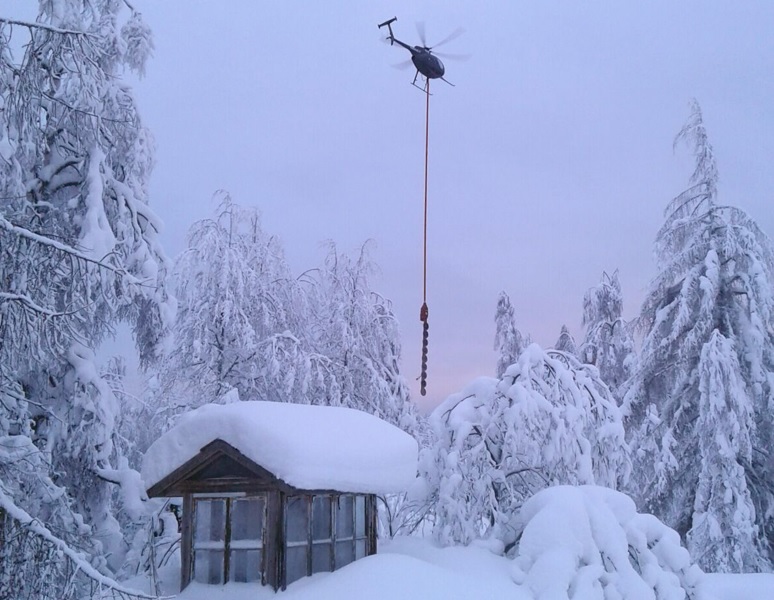 Helikopteri lentää lumisessä maisemassa roikottaen työkalua, joka pudottaa tykkylunta puista.