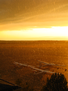 Aurinko paistaa ja sataa lentokentällä. Purjelentokoneita kentällä.