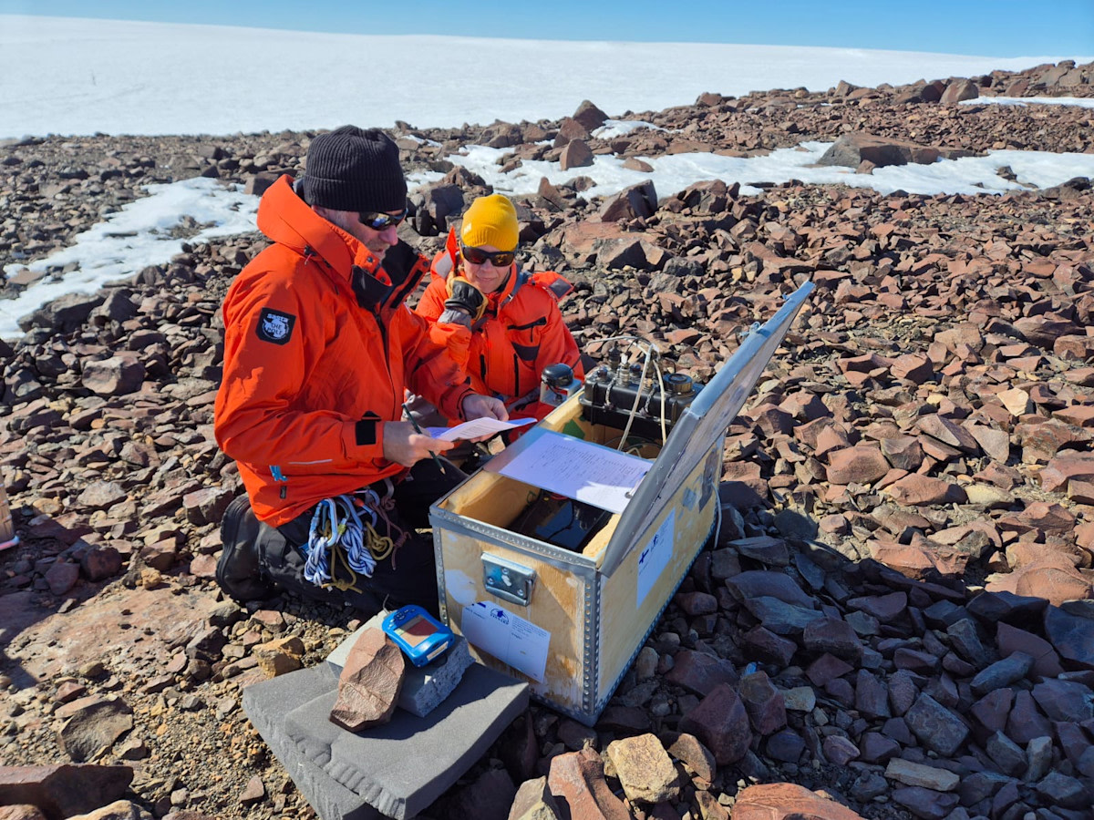 Toppavaattein ja aurinkolasein varustautuneet tutkijat kivikossa mittalaitteen kanssa, taustalla jäätikkö.