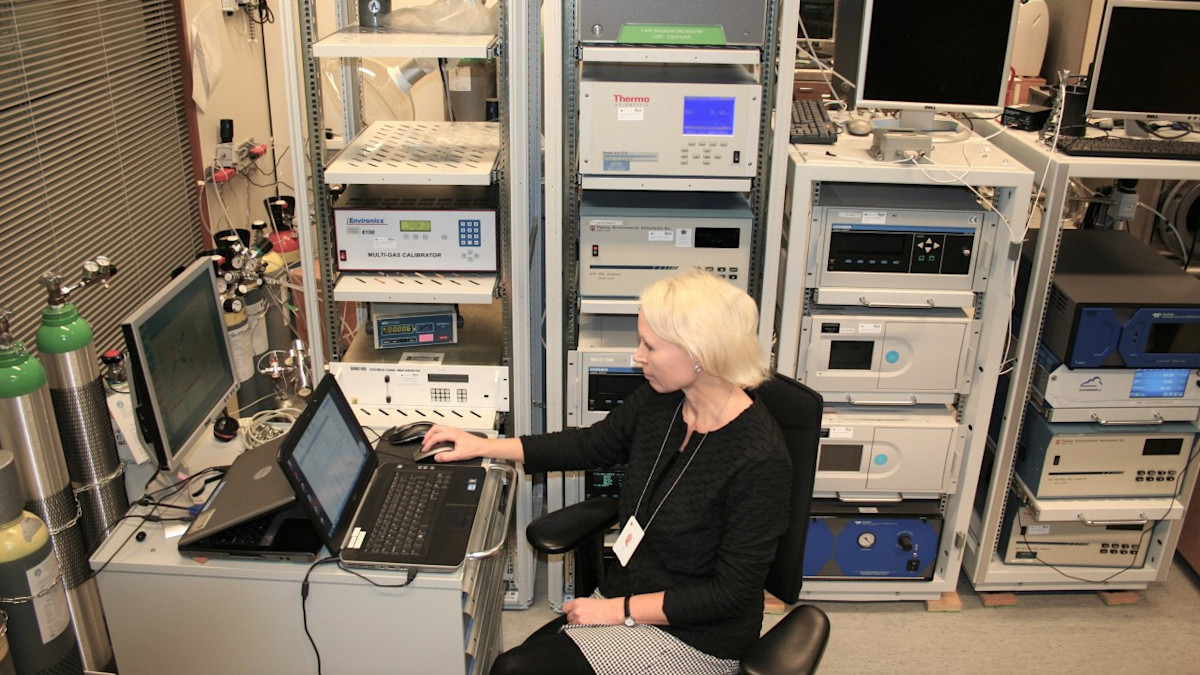 Henkilö istuu tietokoneen ääressä, taustalla laiteräkki.